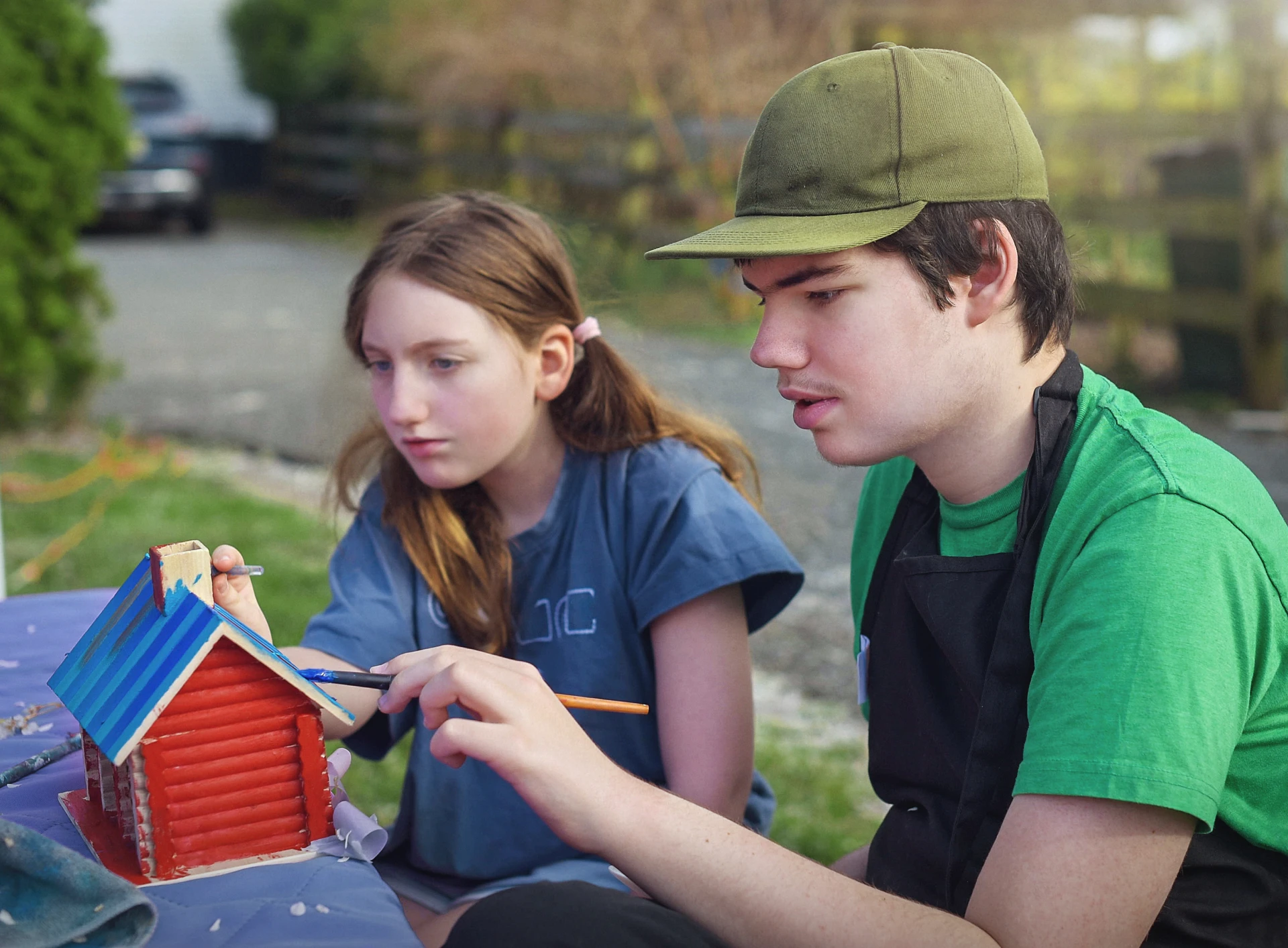 Teens painting a small wooden house outdoors.