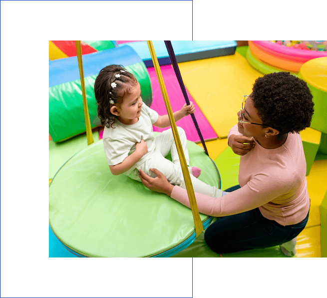 A woman assisting a toddler on playground equipment inside a colorful play area.
